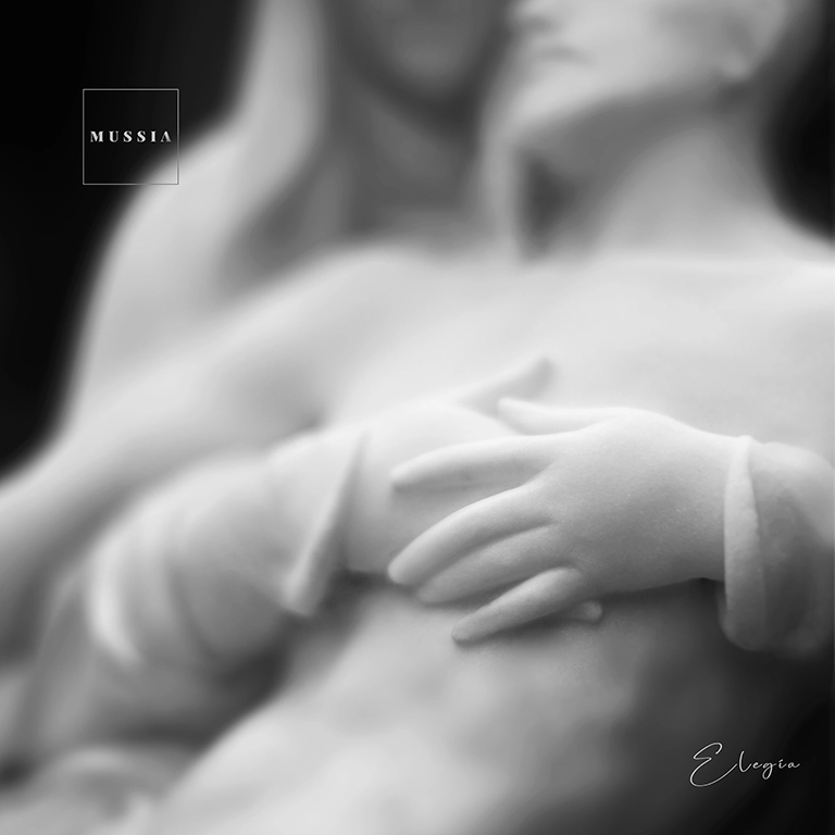 In a white marble sculpture, a woman embraces the chest of her deceased husband while resting him against her chest. "Elegía" Mussia song photo cover. Photo by Jordi Roca Zanuy.