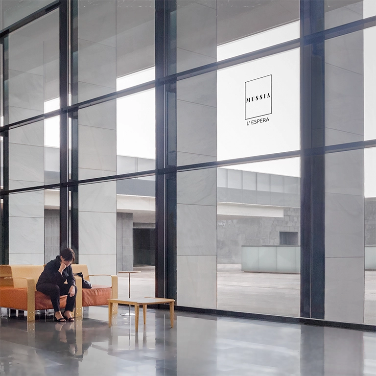 A woman sitting in a large and empty waiting room looks at the floor thoughtfully. "L'Espera" Mussia song photo cover. Photo by Jordi Roca Zanuy.